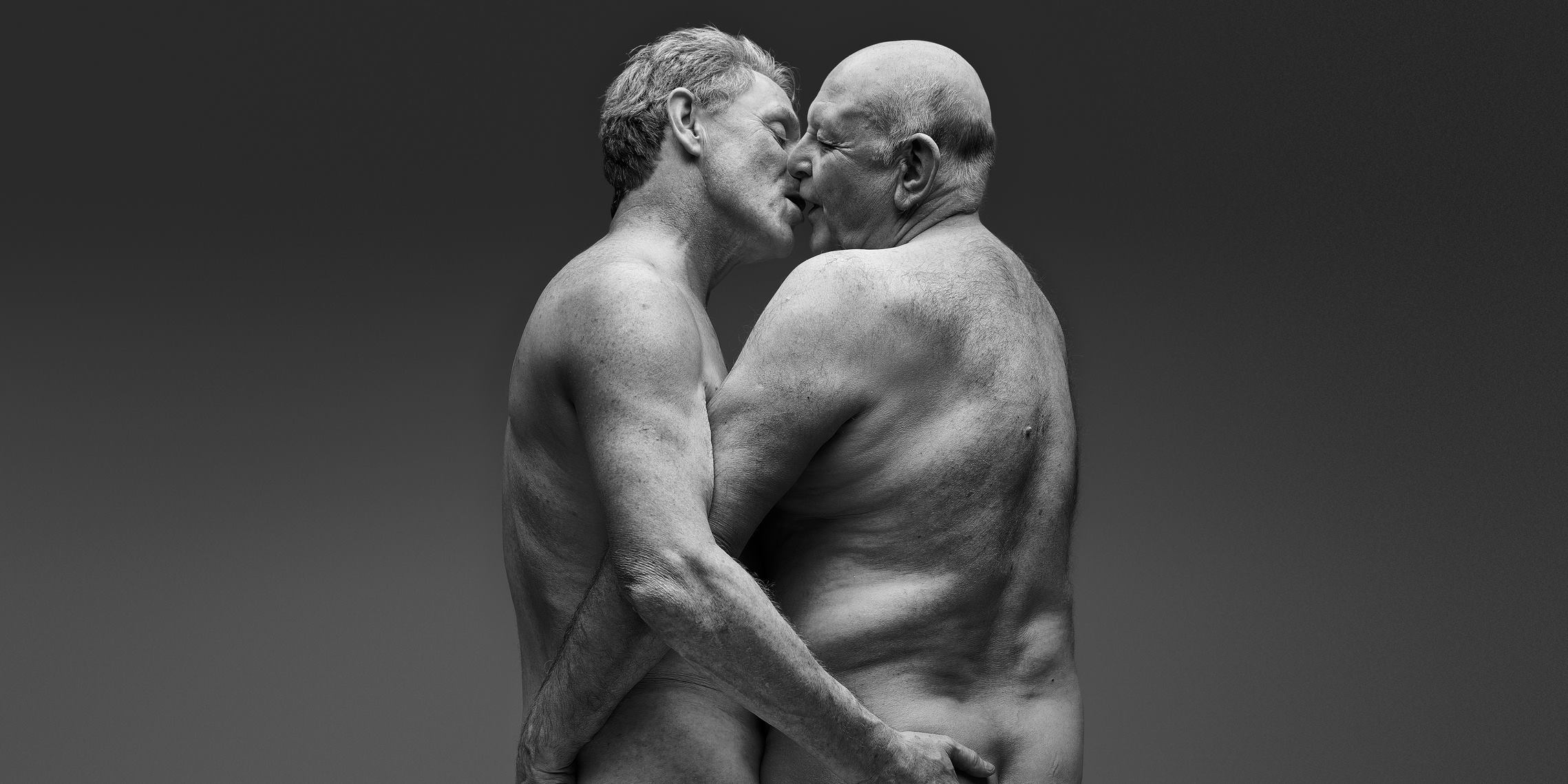 Two men embrace, their mouths are just touching. They hold their hands on each other's naked lower backs, which disappear out of frame but suggest complete nudity