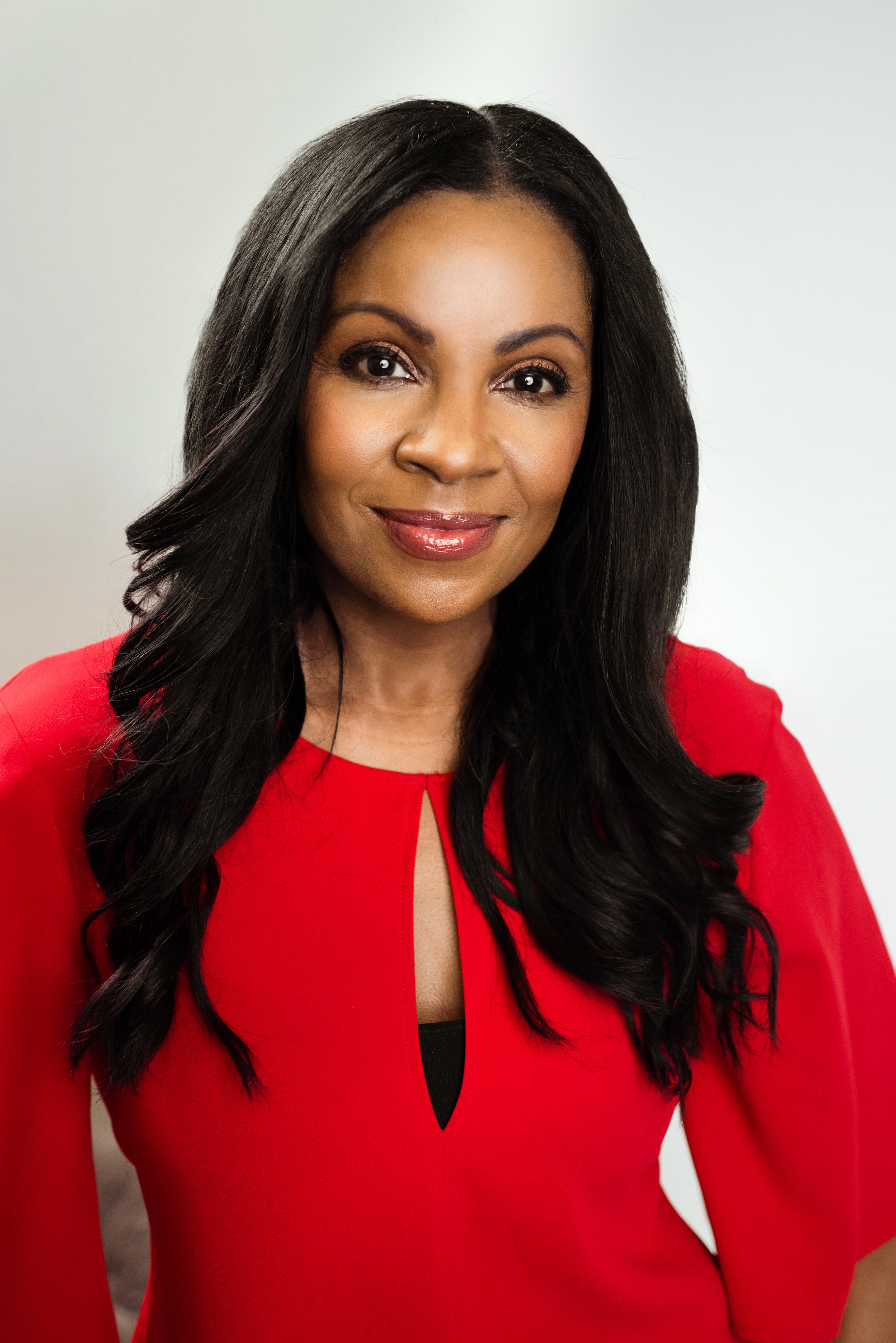A photo of Anjula Mutanda, who has brown, clear skin, wearing a bright red top and looking directly into the camera