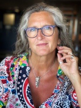 A photo of Dr. Jacqui Gabb, a white woman with long, flowing white and grey hair. She is wearing blue glasses and a brightly patterned top. Her hand is moving towards her chin as if she is about to lean on it.
