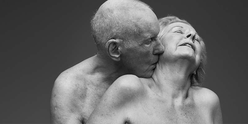 An old man stands behind an old woman, kissing her neck while she closes her eyes. Both are naked.