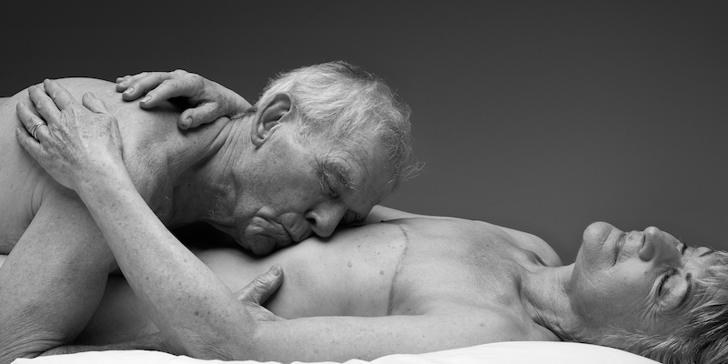 An older woman lies on her back with her eyes closed, mastectomy scars are visible. Her arms are loosely wrapped around an older man who lies on top of her, his head resting on her naked stomach.