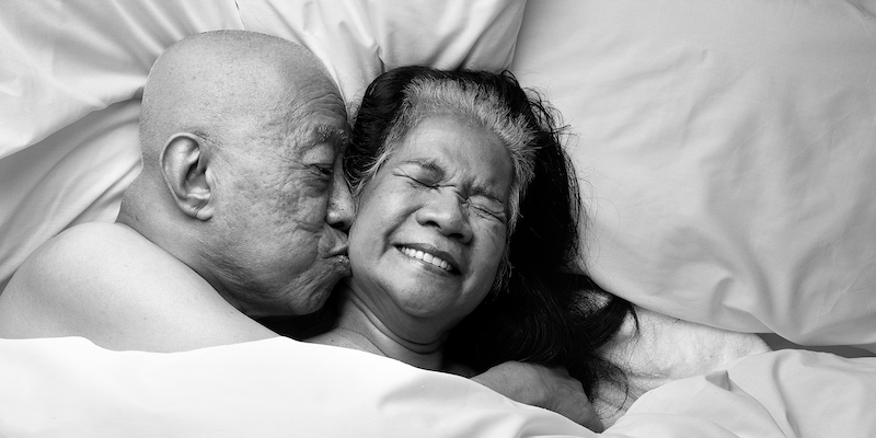 A couple lie in bed together. It's hinted that they're naked but the covers are pulled right up. One is on the left, lying on his side kissing the cheek of his partner, who is facing towards the camera, eyes closed, grinning with delight