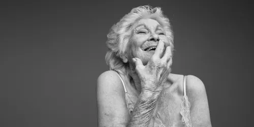 A photo of an older person, the top of lingerie visible as they run their hand down their face smiling